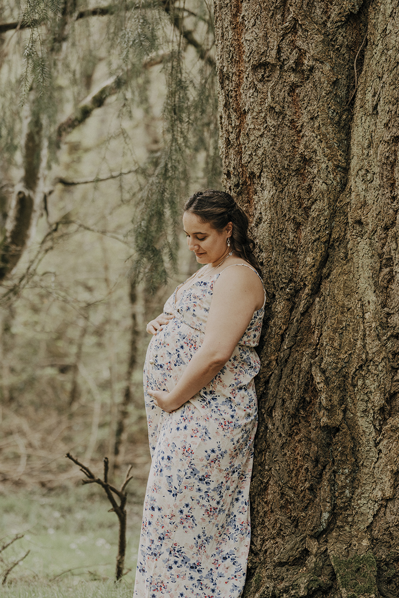 Une future maman qui pose devant un arbre tenant son ventre bien rond dans une robe fleurie